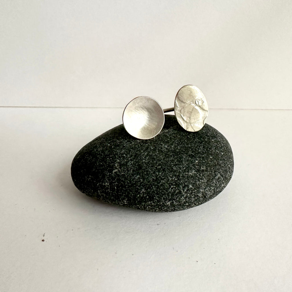 Side by side open silver ring - circle and textured oval shapes sitting on a dark pebble. www.wyckoffsmith.com