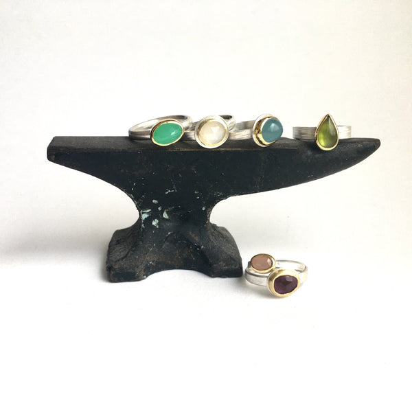 Assortment faceted gemstone silver and gold stacking rings on a mini jeweller's anvil by Michele Wyckoff Smith - left to right: chrysoprase, white moonstone, aquamarine, idiocrase, peach moonstone and pink tourmaline